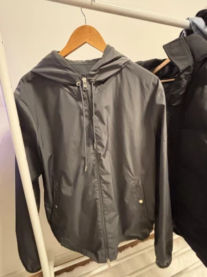 Grå vindjacka med huva och ränder - Grå vindjacka med huva och dragkedja framtill. Jackan har snörning vid halsen och två fickor med knappstängning. På huvan finns en snygg detalj med röd, vit och blå rand. Perfekt för dig som gillar stilrena och sportiga plagg.