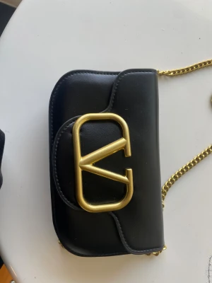 Svart Valentino axelväska med guldkedja - Snygg svart axelväska från Valentino med ett stort guldfärgat V-logo på framsidan. Väskan har en stilren, rundad form och en chunky guldkedja som axelrem. 