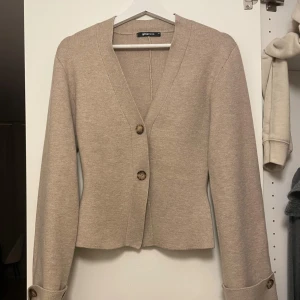 Beige kofta från Gina Tricot - Stilren beige kofta från Gina Tricot med v-ringning och två stora knappar framtill. Koftan är stickad och har långa ärmar med dekorativa knappar vid ärmsluten. Perfekt för lager-på-lager och enkel att matcha med allt.
