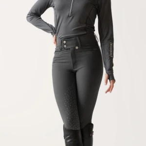 Mörkgrå ridbyxor  - Snygga mörkgrå ridbyxor från Pomme med hög midja, två knappar fram och praktiska fickor. Silikonprint på insidan av benen och bak ger extra grepp i sadeln, endast knee grip. Stretchigt syntetmaterial för skön passform och rörelsefrihet. Perfekta för dig som älskar ridning.