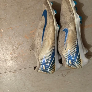 Nike Mercurial Air Zoom fotbollsskor - Nike Mercurial Air Zoom fotbollsskor i vitt och blått med snygga grafiska detaljer på sidan och hälen. Skorna har en låg profil, platt sula med dobbar och är tillverkade i syntetmaterial med ventilerande mesh. Perfekt för dig som vill ha grymt grepp och lätt känsla på planen.