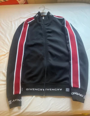 Svart bomberjacka Givenchy S - Givenchy bomberjacka i svart med röda och vita ränder längs ärmarna. Jackan har dragkedja framtill, ribbade muddar och Givenchy-logga på krage och nederkant. Snygg, sportig look med normal passform och lång ärm. Perfekt för dig som gillar streetwear och designer.