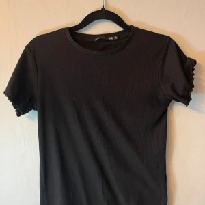 Svart ribbad t-shirt med volangärm - Svart t-shirt från H&M med ribbad struktur och korta ärmar. Ärmsluten har en söt volangdetalj som ger en extra touch. Rund halsringning och mjukt material som känns skönt mot huden. Perfekt för dig som gillar enkel men snygg stil. Från lager157 i strl S
