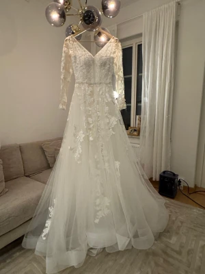 Vit långklänning med spets och slöja - Drömmig vit långklänning med v-ringad överdel, långa spetsärmar och blommiga spetsdetaljer. Kjolen består av flera lager tunn tyll som ger extra volym och vackert flow. En fluffig underkjol, lång slöja i tunn tyll och ett glittrigt diadem ingår. Originalpris: 25 000 kr. 