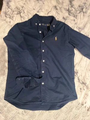 Mörkblå skjorta från Ralph Lauren - Snygg mörkblå skjorta från Ralph Lauren med klassisk button-down krage och broderad logga på bröstet. Skjortan har vita knappar och långa ärmar, perfekt för dig som gillar stilrena och tidlösa plagg. Materialet är mjuk bomull som känns skönt mot huden.