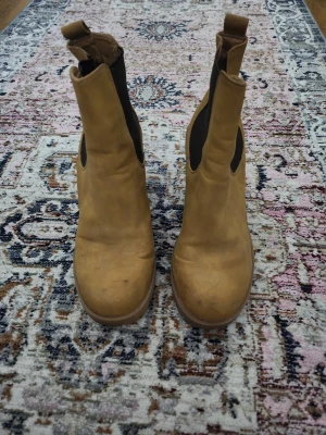 Beige chelsea boots med klack -  Beige chelsea boots med bred klack och rund tå. Skorna har elastiska svarta paneler på sidorna och är tillverkade i mjukt skinn.