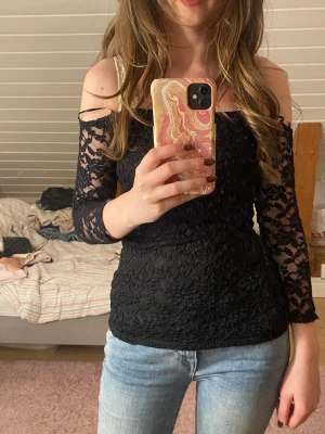 Svart spetsig offshoulder topp - Säljer en svart offshoulder topp med långärmade spetsdetaljer. Toppen har en tight passform och är tillverkad i ett snyggt spetsmaterial som ger en cool och elegant vibe. Perfekt att styla med jeans eller kjol för en trendig look.