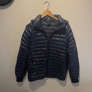 Mörkblå pufferjacka från Peak Performance storlek (170) - Snygg mörkblå pufferjacka från Peak Performance med huva och svarta detaljer. Jackan har quiltad design, dragkedja framtill och två sidofickor med dragkedja. Peak Performance-logga på ärmen och på huvan. Perfekt för kyliga dagar.