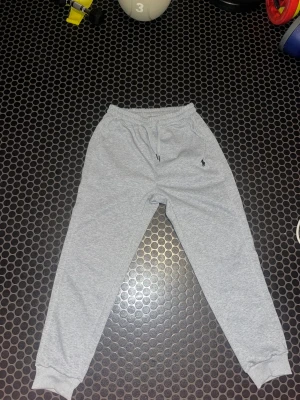 Grå mjukisbyxor från Polo Ralph Lauren - Snygga grå mjukisbyxor från Polo Ralph Lauren med klassisk logga broderad på benet. Skulle säga att byxorna passar mer för S/M Byxorna har elastisk midja med snörning, ribbade muddar vid benslut och är gjorda i mjukt bomullsmaterial. Perfekta för chill eller träning.