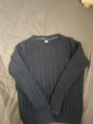 Mörkblå kabelstickad tröja från CRW - Kabelstickad mörkblå tröja från CRW med rund halsringning och långa ärmar. Tröjan har ett klassiskt flätmönster och ribbade muddar vid ärmslut och nederkant. Perfekt för lager-på-lager under kyliga dagar.