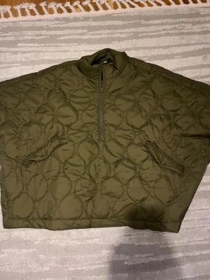 Olivgrön quiltad pufferjacka - Säljer en oversized olivgrön pufferjacka med quiltat mönster och half zip framtill. Jackan har höga krage, två fickor med knappstängning och är tillverkad i syntetmaterial. Perfekt för dig som gillar chill och loose fit. Västen/jackan är aldrig använd och är perfekt till hösten o vintern. 