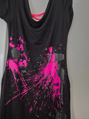 Klänning - Svart kortklänning med coolt neonrosa och grått splattermönster framtill. Klänningen har korta ärmar, rundad hals och snygga neonrosa band i ryggen som ger en edgy vibe. Mjuk och stretchig bomullskvalitet, perfekt för dig som gillar att sticka ut.