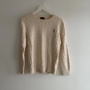 Beige kabelstickad tröja Polo Ralph Lauren - Kabelstickad tröja från Polo Ralph Lauren i beige med rund hals och lång ärm. Klassisk design med ribbade muddar och diskret broderad logga på bröstet. Perfekt för dig som gillar stilrena och tidlösa plagg. Obs att detta är en herrtröja. 