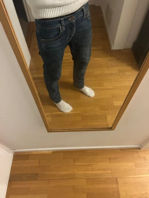 Blå jeans från Nudie Jeans Co - Säljer ett par snygga blå jeans från Nudie Jeans Co med klassisk femficksdesign och orangea sömmar. Jeansen har en normal passform och raka ben, med tydliga slitningar för en cool look. Perfekt för dig som gillar stilrena och tidlösa jeans.