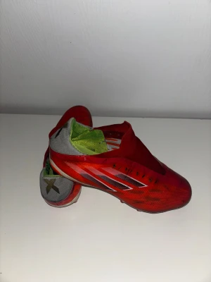 Röda Adidas fotbollsskor - Säljer ett par röda Adidas fotbollsskor med silverfärgade detaljer och grå häl. Skorna har en strömlinjeformad design utan synlig snörning och insida i limegrönt. Yttermaterialet är syntet och sulan har dobbar för gräsplan. Perfekt för dig som vill sticka ut på planen. Pris kan diskuteras 