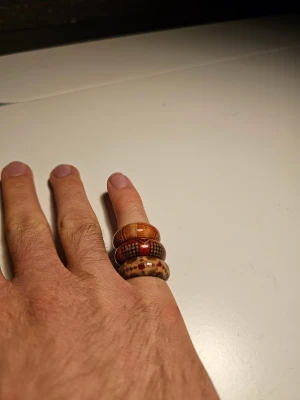 Tre coola mönstrade ringar i trä - Säljer ett set med tre unika ringar i trä, alla med olika mönster och färger. En är brun med rutigt mönster, en är rödbrun med prickar och en är ljusbrun med små röda detaljer. Alla är I trä och ca 19 mm