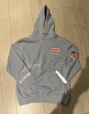 Ljusgrå NASA hoodie med tryck - Säljer en ljusgrå hoodie från NASA med röd logga på bröstet och amerikansk flagga på ärmen. Tröjan har huva med snörning, magficka och vita detaljer på ärmar och rygg. Materialet är mjukt och känns som polyester. Prislapp sitter kvar