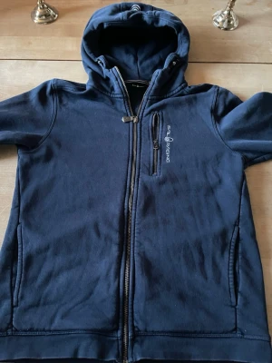 Marinblå hoodie från Sail Racing - Snygg marinblå hoodie från Sail Racing med dragkedja framtill, huva och tre fickor med dragkedja. Broderad logga på bröstet och mjukt material som känns skönt mot huden. Perfekt för en avslappnad och sportig stil. 14 år /164cm lång 
