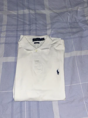 Vit pikétröja från Polo Ralph Lauren - Klassisk vit pikétröja från Polo Ralph Lauren med korta ärmar och blå broderad logga på bröstet. Tröjan har krage och knappar vid halsen. Tillverkad i mjuk bomull för en skön och stilren look.