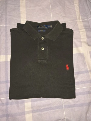 Svart pikétröja från Polo Ralph Lauren - Klassisk svart pikétröja från Polo Ralph Lauren med röd broderad logga på bröstet. Tröjan har krage, två knappar framtill och är tillverkad i mjuk bomull. Perfekt till en avslappnad men ändå stilren look.