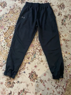 Svarta mjukisbyxor från Under Armour - Svarta mjukisbyxor från Under Armour med resår i midjan och muddar vid bensluten. Byxorna har en diskret logga på ena låret och texten UNDER ARMOUR längs ena benet. Perfekta för chill eller träning, tillverkade i ett lätt och bekvämt material.