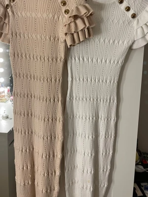 Stickad klänning med volangärmar - Två stickade klänningar i beige och vitt med dekorativa guldknappar vid axlarna och volangärmar. Klänningarna har ett strukturerat mönster och är figurnära i modellen. Perfekt för dig som gillar trendiga detaljer och stickat. Köpte för 1000kr st från raglady säljer för 700kr/st