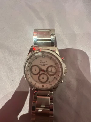Silverfärgad klocka från Regal - Stilren klocka från Regal med silverfärgat armband i metall och vit urtavla. Klockan har flera små urtavlor och tydliga siffror runt om. Perfekt accessoar för dig som gillar en klassisk look. Funkar ej!