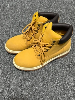 Gula kängor med svart kant - Säljer ett par snygga gula kängor med svart vadderad kant och vita sulor. Kängorna har klassisk snörning och robust design, perfekta för dig som gillar streetstyle. Materialet ser ut att vara läder eller mocka och de har en cool urban vibe.