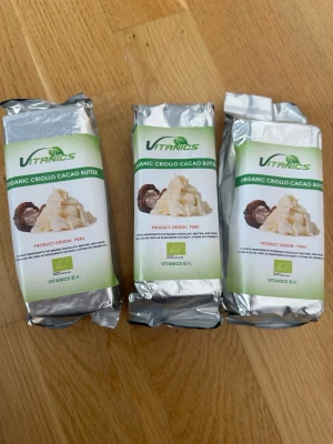 Ekologiskt Criollo kakaosmör 3x250g - Tre paket ekologiskt Criollo kakaosmör från Vitanics, ursprung Peru. Varje paket väger 250g och är inslaget i silverfärgad folie med grön och vit etikett. Perfekt för bakning, hudvård eller raw food-recept.