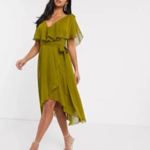 Olivgrön omlottklänning med volang - Säljer en olivgrön omlottklänning med v-ringning och volangdetaljer. Klänningen har kort ärm, asymmetrisk nederkant och knyt i midjan för en snygg siluett. Perfekt för dig som vill ha en flowy och trendig look.