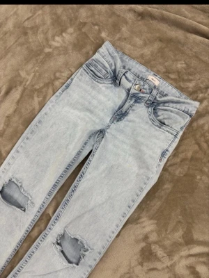 Ljusblå ripped jeans med brodyr - Säljer ett par ljusblå jeans med slitningar på knäna och coola röda broderade detaljer på bakfickorna. Jeansen har klassisk femficksmodell och är i stretchigt denimtyg. Perfekta för en avslappnad och trendig look.