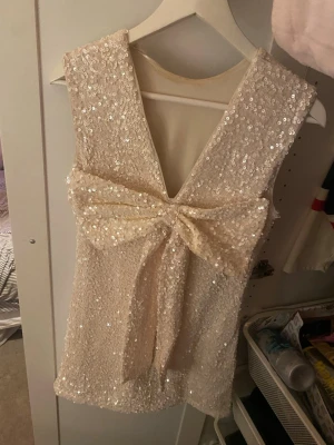 Glittrig paljettklänning med rosett - Supersnygg ärmlös klänning i ljusbeige med massor av paljetter och en stor rosett i ryggen. Djup v-ringning bak och figurnära passform. Perfekt för dig som vill sticka ut och glänsa på nästa event!