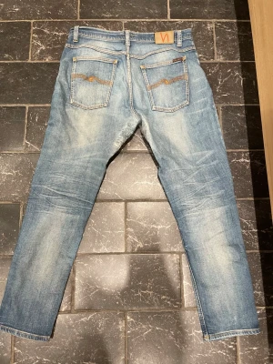 Ljusblå jeans från Nudie Jeans - Säljer ett par ljusblå jeans från Nudie Jeans med klassisk femficksmodell och raka ben. Jeansen har slitningar och snygg tvätt, samt kontrastsömmar och Nudie-logga på bakfickan. Tillverkade i bomull för en skön känsla. Lagade i grenen!!!! Storlek 33/30 men skulle uppskatta 33/32