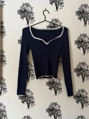 Mörkblå ribbad långärmad topp - Trendig mörkblå ribbad topp med lång ärm och ljusgrå kant vid halsringning och nederkant. Toppen har en snygg v-ringning och croppad passform som ger en modern look. Perfekt att styla med höga jeans eller kjol.