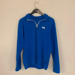 Blå halvzip tröja från Helly Hansen - Snygg blå långärmad tröja från Helly Hansen med sopren design och vit HH-logga på bröstet. Tröjan har halv dragkedja framtill och hög krage och en sportig look.