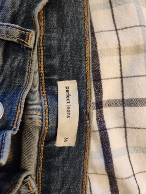 Perfect Jeans från Gina Tricot - Säljer ett par klassiska blå jeans från Perfect Jeans i storlek 34. Jeansen har en snygg tvätt, kontrastsömmar och dragkedja med knapp framtill. Köpta på Vinted men dem passade inte. Innerbenslängden är 70cm. Inga defekter vad jag har sätt. Men jag har dock bara privat ena benet ich sen har dem hängt i garderoben.