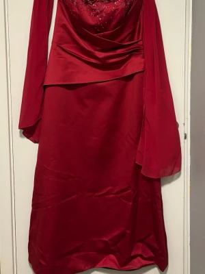 Röd långklänning/Balklänning  - Elegant röd långklänning utan axelband eller ärmar och draperad överdel. Klänningen har dekorativa detaljer upptill och är tillverkad i ett glansigt material som ger en lyxig känsla. Perfekt för dig som vill sticka ut med en klassisk och sofistikerad look. Passar som balklänning. 