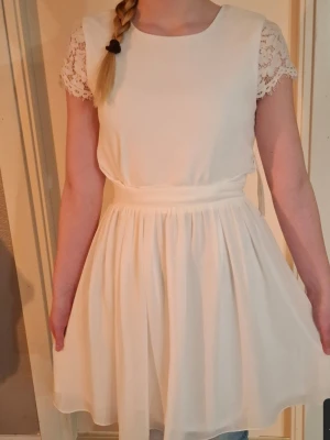 Vit kortklänning med spetsärmar - Söt vit klänning med kort längd och spetsdetaljer på ärmarna. Klänningen har rund halsringning, markerad midja och ett lätt, flödande kjolparti. Perfekt för somriga dagar eller speciella tillfällen. Ny med lappar kvar