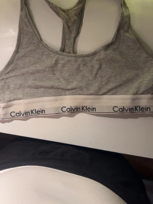Grå sport-bh från Calvin Klein -  En grå sport-bh från Calvin Klein med klassisk brottarrygg och bred resår med logga runt om. Bh:n är tillverkad i mjukt och stretchigt material, perfekt för en avslappnad och sportig look. Använd ett antal gånger och inget fel med den förutom defekterna som syns i bilden, bleknings fläckar från hår färg som ej går bort men annars bra skick! Storlek S och säljer pågrund av att den inte kommer till användning💕