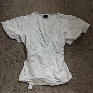 Ljusblå omlottopp med knytband - Supersnygg ljusblå omlottopp från Monki med djup v-ringning och breda korta ärmar. Toppen har knytband i midjan som ger en fin siluett och är tillverkad i mjuk bomull. Perfekt för dig som vill ha en chill men ändå stilren look.