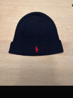 Mörkblå mössa från Polo Ralph Lauren - Snygg mörkblå mössa från Polo Ralph Lauren i mjuk merinoull. Klassisk ribbstickad kant och ikonisk röd logga broderad framtill. Perfekt accessoar för att hålla värmen och lyfta din stil under kyliga dagar.