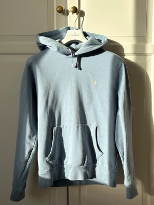 Ljusblå hoodie från Polo Ralph Lauren - Ljusblå hoodie från Polo Ralph Lauren med klassisk huva och känguruficka framtill. Liten broderad logga i vitt på bröstet. Tillverkad i mjukt bomullsmaterial och har ribbade muddar vid ärmslut och nederkant. Perfekt för en avslappnad och stilren look.