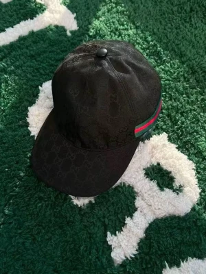 Grå Gucci keps med orm och GG-mönster - Säljer en grå keps från Gucci med klassiskt GG-mönster och en stor röd, vit och blå orm broderad på framsidan. Kepsen har svart mesh baktill och justerbar rem. Märkesdetaljer både på knapp och insida. Perfekt för dig som vill sticka ut.