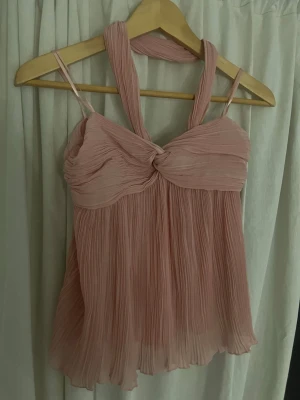 Plisserad rosa linnetopp från Zara  - Supersöt plisserad rosa linnetopp från H&M med twistad detalj framtill och tunna axelband. Toppen har en flowy passform och är perfekt för dig som vill ha en feminin och trendig look. Materialet är lätt och luftigt, vilket gör den skön att bära under varma dagar.