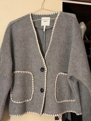 Grå cardiganjacka från OBJECT - Grå cardiganjacka från OBJECT med två stora fickor framtill och dekorativa vita kanter. Jackan har V-ringning, två svarta knappar och är gjord i ett mjukt, ull-liknande material. Perfekt för lager på lager och en chill look.