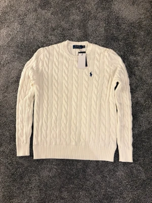 Vit kabelstickad tröja Polo Ralph Lauren - Kabelstickad vit tröja från Polo Ralph Lauren med rund halsringning och broderad marinblå logga på bröstet. Tröjan har långa ärmar och ribbade muddar vid ärmslut och nederkant. Perfekt för en stilren och klassisk look.
