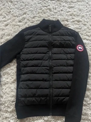 Svart cardiganjacka från Canada Goose - Snygg svart cardiganjacka från Canada Goose med quiltad front och stickade ärmar. Jackan har dragkedja framtill och en rund halsringning. Märkets logga sitter på vänster ärm. Perfekt för lager-på-lager och streetwear. Höger dragkedja till fickan är trasig annars i gott skick
