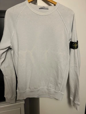 Ljusgrå sweatshirt från Stone Island - Ljusblå sweatshirt från Stone Island med klassisk rund halsringning. Tröjan har den ikoniska svarta logotypen med kompass på vänster ärm. Perfekt för en avslappnad och stilren look. Storlek S