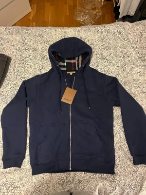 Marinblå zip hoodie från Burberry - Snygg marinblå hoodie från Burberry med dragkedja framtill och klassiskt rutigt foder i huvan. Hoodien har två fickor fram, ribbade muddar och justerbar dragsko i huvan. Perfekt för en chill och stilren look.