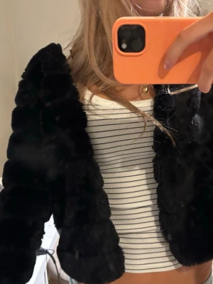 Svart fluffig pälsjacka - Säljer en svart, kort pälsjacka med fluffig och mjuk känsla. Jackan har en rundad krage och långa ärmar, perfekt för att hålla värmen under kalla dagar. Passar dig som vill ha en trendig och mysig look till vintern. 